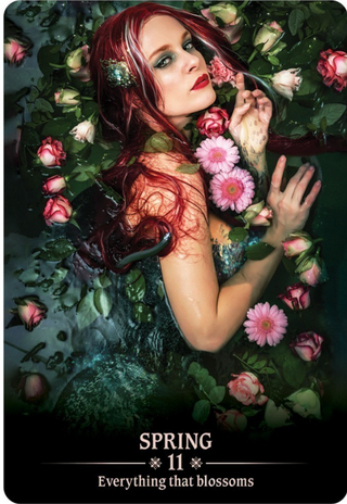 The Spring card from the Dark Light oracle deck shows a woman with red hair lying in a pool of water, surrounded by pink flowers. 